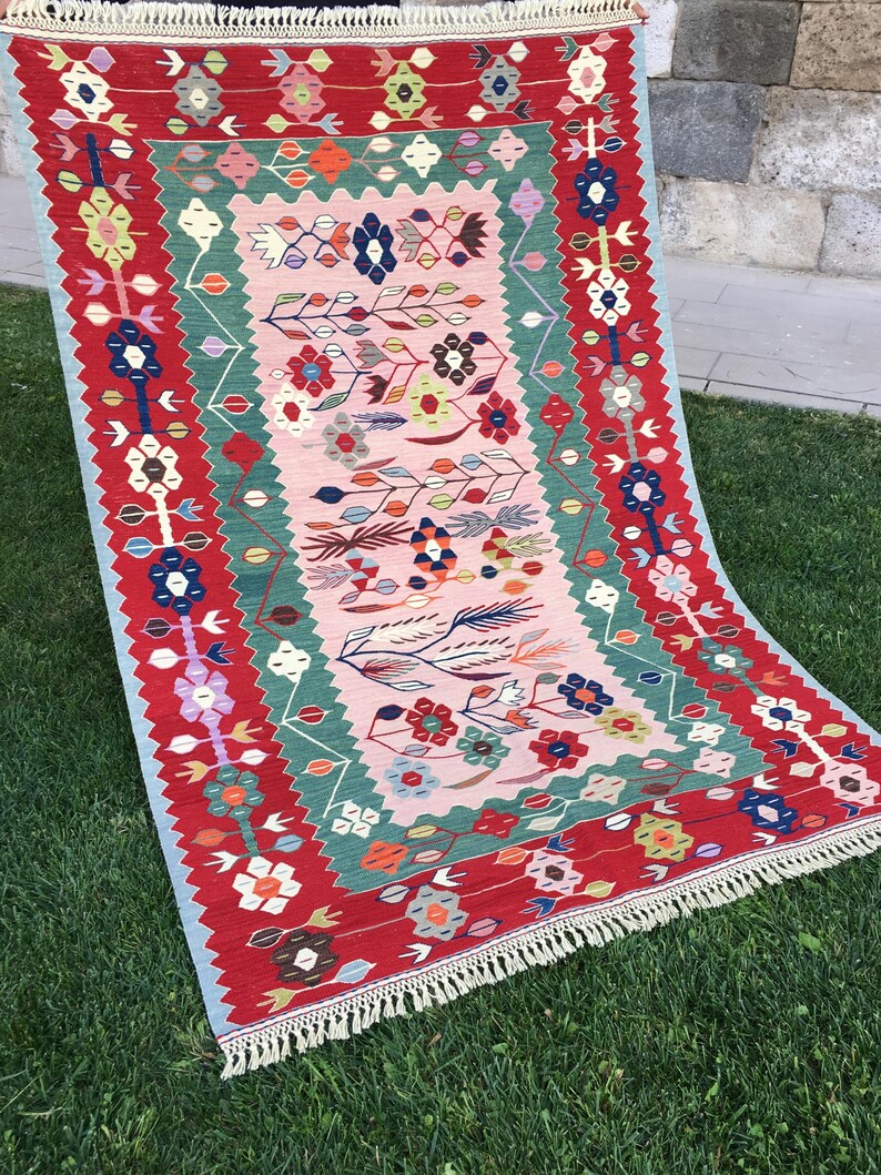 May include: A rectangular, hand-woven rug with a floral design. The rug features a red border with colorful floral motifs, a central pink field, and green and blue accents. The rug has a fringed edge and is displayed on green grass.