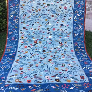 May include: A rectangular, hand-woven rug with a light blue central field and a dark blue border. The rug features a floral design with various colored flowers and leaves. The border has a repeating floral pattern, and the rug has a fringe along the bottom edge.