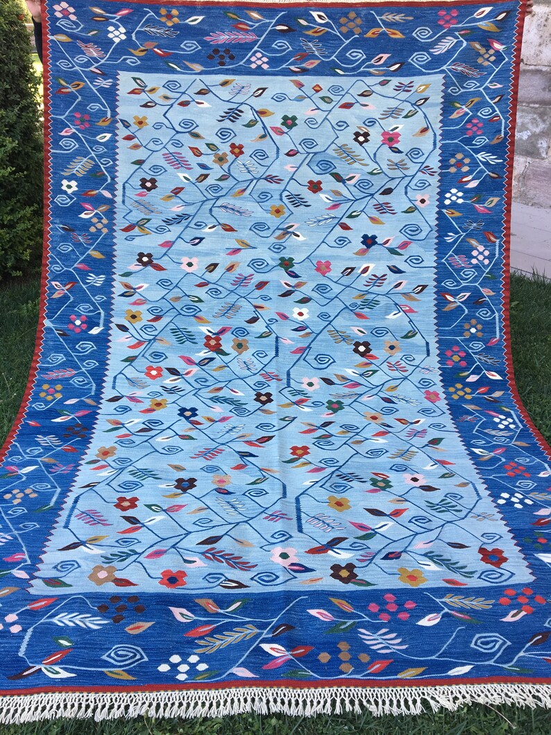 May include: A rectangular, hand-woven rug with a light blue central field and a dark blue border. The rug features a floral design with various colored flowers and leaves. The border has a repeating floral pattern, and the rug has a fringe along the bottom edge.