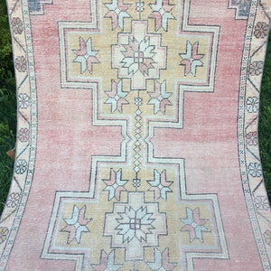 May include: A vintage rug with a central geometric design in shades of peach, beige, and gray. The rug features a repeating pattern of stylized floral and geometric shapes, with a border of smaller floral motifs. The rug's distressed appearance adds to its charm.