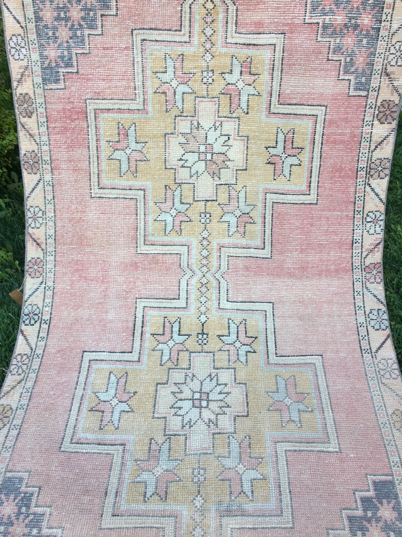 May include: A vintage rug with a central geometric design in shades of peach, beige, and gray. The rug features a repeating pattern of stylized floral and geometric shapes, with a border of smaller floral motifs. The rug's distressed appearance adds to its charm.