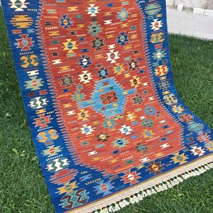 Puede incluir: Una alfombra turca tradicional con fondo azul y rojo. La alfombra tiene un patr&oacute;n geom&eacute;trico con acentos blancos, amarillos y verdes. La alfombra tiene flecos en la parte inferior.