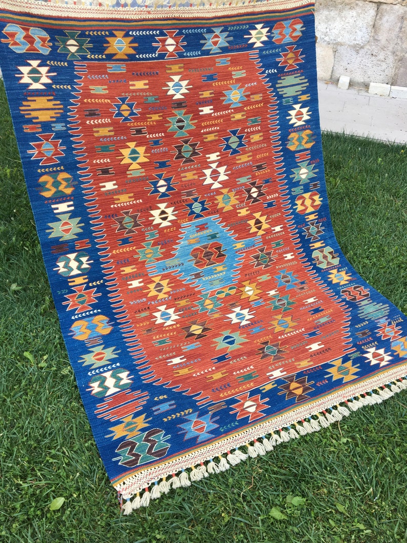 Puede incluir: Una alfombra turca tradicional con fondo azul y rojo. La alfombra tiene un patr&oacute;n geom&eacute;trico con acentos blancos, amarillos y verdes. La alfombra tiene flecos en la parte inferior.