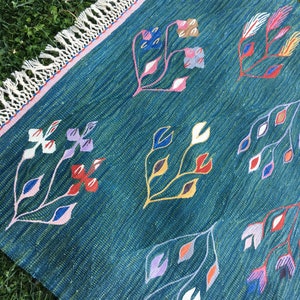 Puede incluir: Una alfombra azul turquesa con borde de flecos y un patr&oacute;n floral en tonos de rosa, rojo, amarillo, azul y blanco.