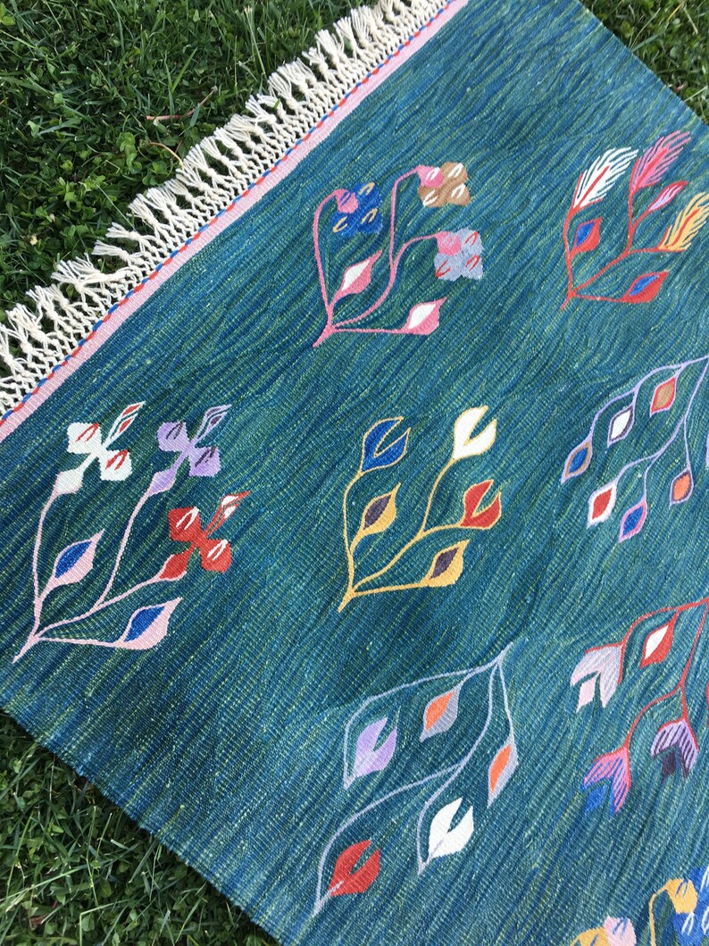 Puede incluir: Una alfombra azul turquesa con borde de flecos y un patr&oacute;n floral en tonos de rosa, rojo, amarillo, azul y blanco.