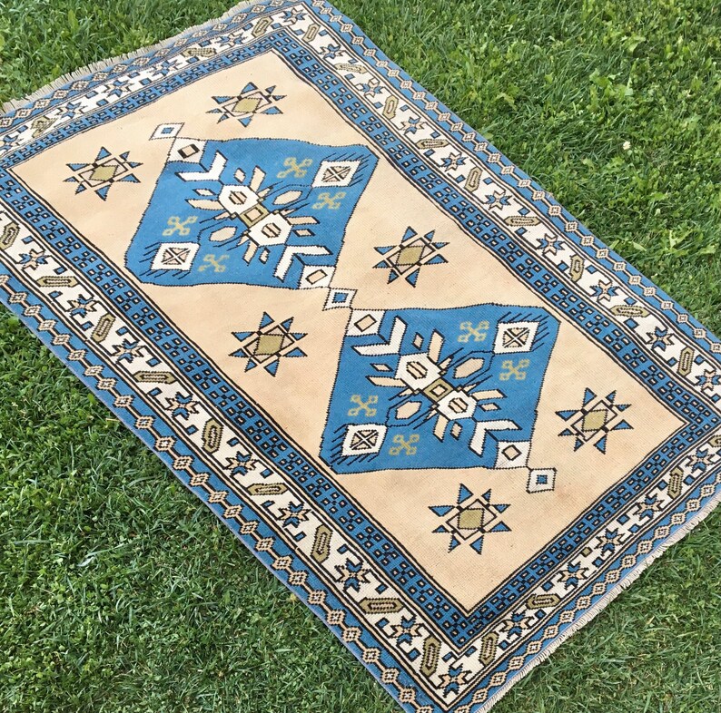 May include: A beige rug with a blue and green geometric pattern. The rug has a fringe border.