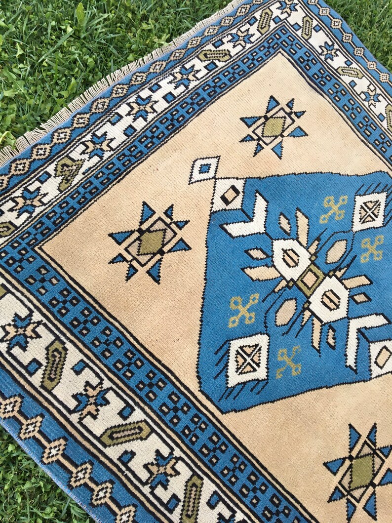 May include: A beige rug with a blue and white geometric pattern. The rug has a fringe border and features a central design with a starburst motif.