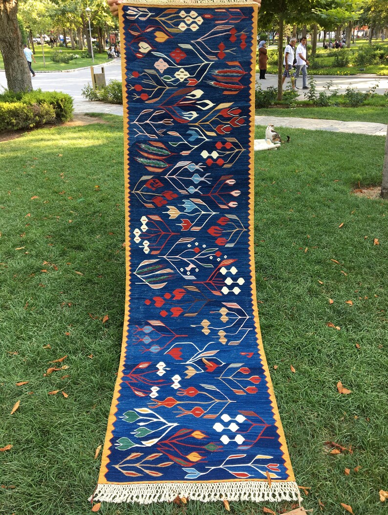 May include: A long, narrow rug with a blue background and a colorful geometric pattern. The rug has a yellow border and white fringe.
