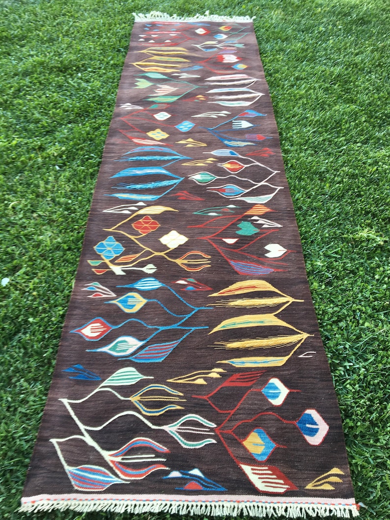 Puede incluir: Una alfombra larga y estrecha con fondo marr&oacute;n y un patr&oacute;n geom&eacute;trico colorido. El patr&oacute;n presenta hojas estilizadas, diamantes y otras formas en tonos de azul, rojo, amarillo, verde y blanco.