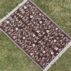 Puede incluir: Una alfombra marr&oacute;n con un patr&oacute;n floral y flecos blancos. La alfombra tiene un dise&ntilde;o geom&eacute;trico con flores y hojas estilizadas.