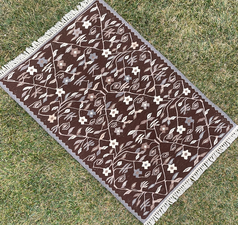 Puede incluir: Una alfombra marr&oacute;n con un patr&oacute;n floral y flecos blancos. La alfombra tiene un dise&ntilde;o geom&eacute;trico con flores y hojas estilizadas.