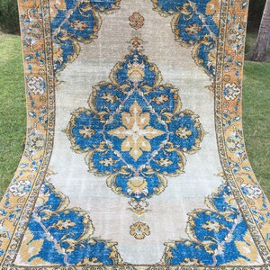 May include: A large, rectangular rug with an ornate, traditional design. The rug features a central medallion and corner elements in shades of blue, gold, and beige. The border is a contrasting gold color. The rug is displayed outdoors on grass.