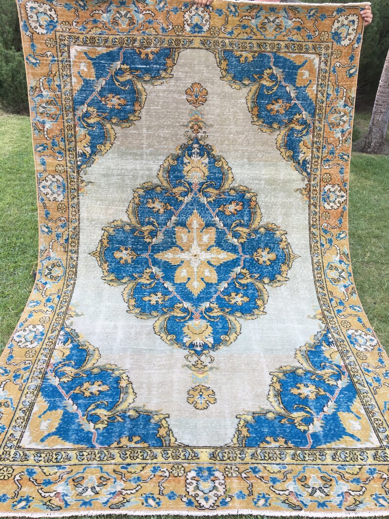 May include: A large, rectangular rug with an ornate, traditional design. The rug features a central medallion and corner elements in shades of blue, gold, and beige. The border is a contrasting gold color. The rug is displayed outdoors on grass.