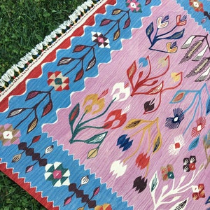 Op de afbeelding: Een kleurrijk tapijt met een roze achtergrond en een blauwe rand. Het tapijt heeft een bloemenpatroon met gestileerde bloemen en bladeren in tinten rood, geel, blauw en groen.
