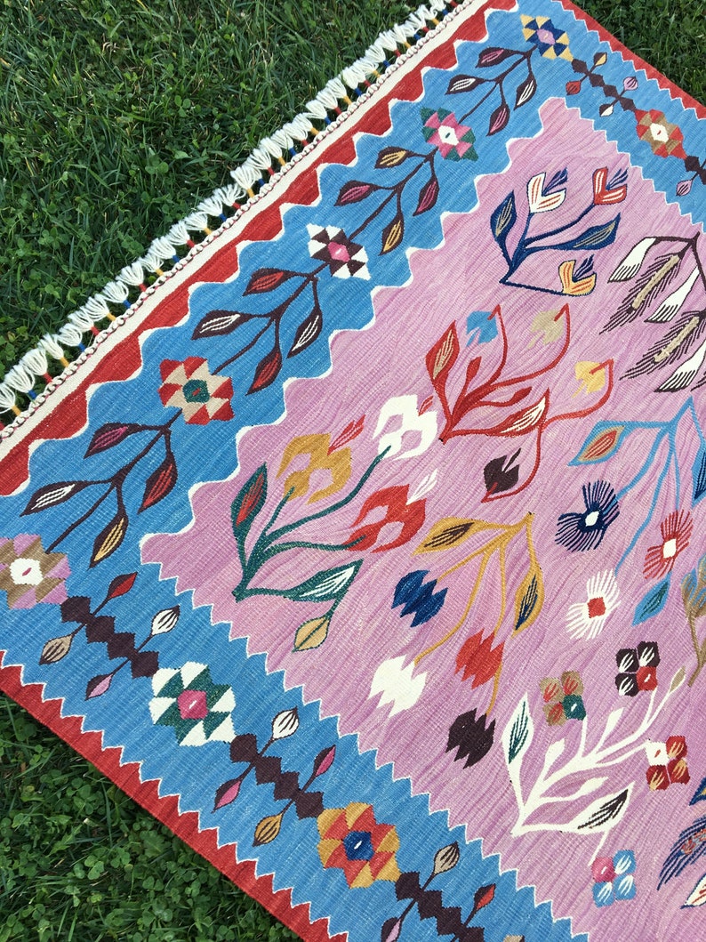 Op de afbeelding: Een kleurrijk tapijt met een roze achtergrond en een blauwe rand. Het tapijt heeft een bloemenpatroon met gestileerde bloemen en bladeren in tinten rood, geel, blauw en groen.