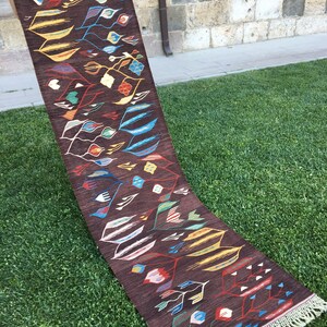 Puede incluir: Una alfombra larga y estrecha con fondo marr&oacute;n y un patr&oacute;n geom&eacute;trico colorido. La alfombra tiene flecos en ambos extremos.