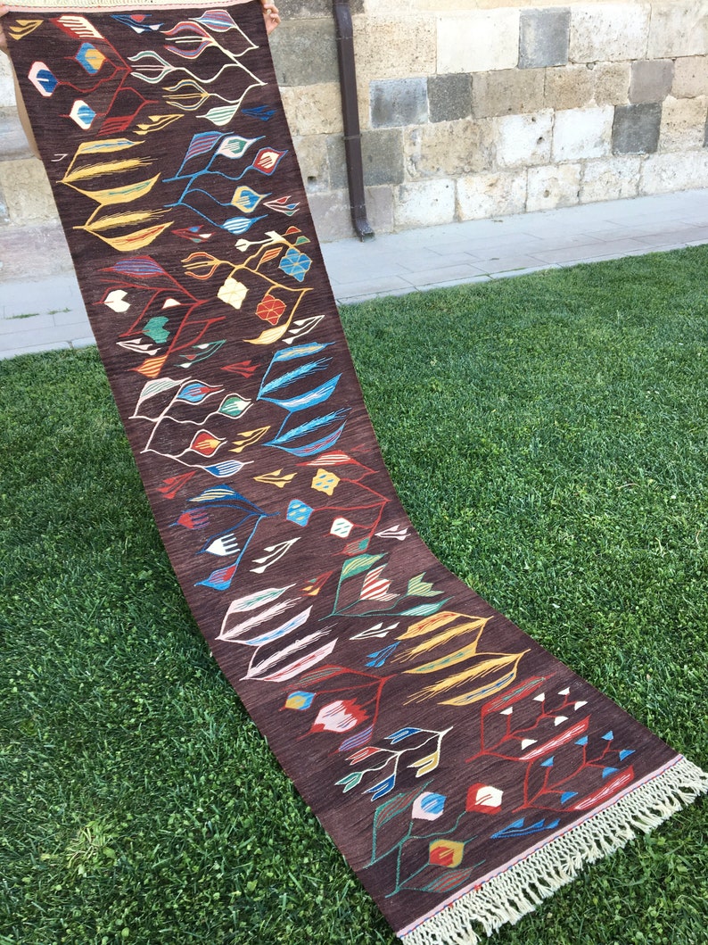 Puede incluir: Una alfombra larga y estrecha con fondo marr&oacute;n y un patr&oacute;n geom&eacute;trico colorido. La alfombra tiene flecos en ambos extremos.