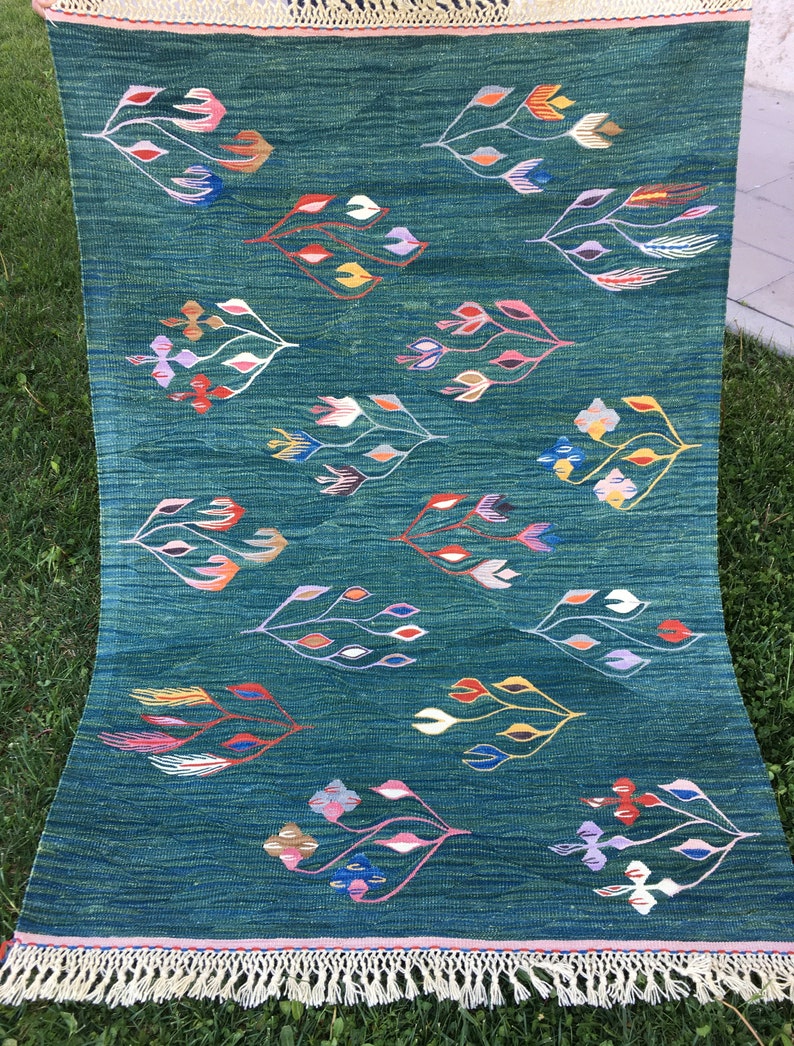 Puede incluir: Una alfombra azul verdosa con borde de flecos presenta un patr&oacute;n repetitivo de dise&ntilde;os florales estilizados en tonos de rosa, naranja, amarillo, azul y verde.
