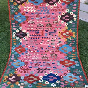 May include: A colourful rug with a pink background and intricate patterns in shades of blue, green, red, and yellow. The rug has a fringed border.