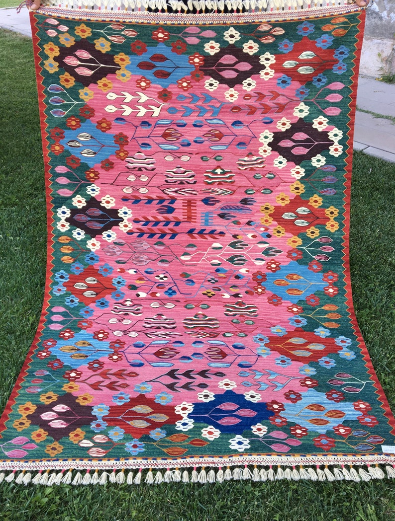 May include: A colourful rug with a pink background and intricate patterns in shades of blue, green, red, and yellow. The rug has a fringed border.