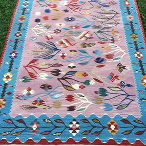 Op de afbeelding: Een kleurrijk tapijt met een roze achtergrond en een blauwe rand. Het tapijt heeft een bloemenpatroon met gestileerde bladeren en bloemen in tinten rood, blauw, geel en groen.