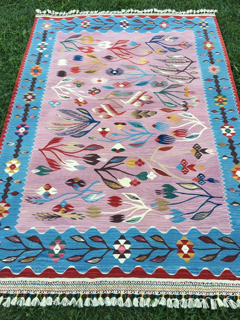 Op de afbeelding: Een kleurrijk tapijt met een roze achtergrond en een blauwe rand. Het tapijt heeft een bloemenpatroon met gestileerde bladeren en bloemen in tinten rood, blauw, geel en groen.