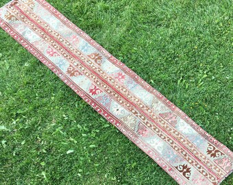 May include: A long, narrow runner rug with a traditional Turkish kilim design. The rug features a repeating pattern of geometric shapes in shades of red, beige, and blue.