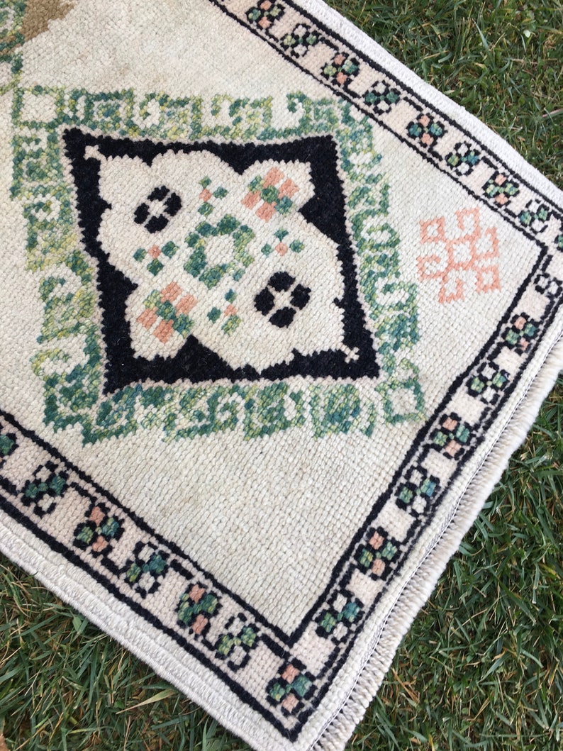 May include: A white rug with a black, green and pink geometric pattern. The rug has a fringed border.
