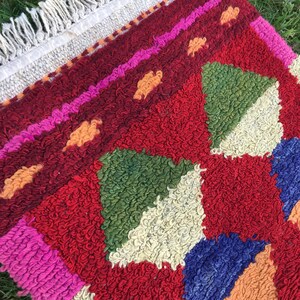 Puede incluir: Una alfombra colorida tejida a mano con un patr&oacute;n geom&eacute;trico en tonos de rojo, rosa, verde, azul y naranja. La alfombra tiene un borde de flecos.