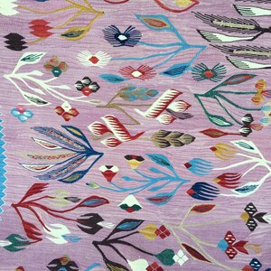 Op de afbeelding: Een roze tapijt met een bloemenpatroon. Het tapijt heeft een verscheidenheid aan kleuren, waaronder rood, blauw, groen en geel. De bloemen zijn gestileerd en abstract, en het tapijt heeft een vintage uitstraling.