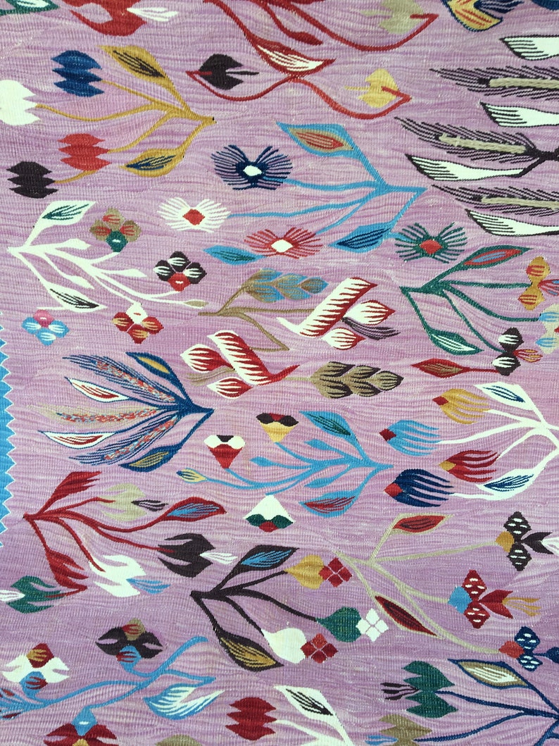 Op de afbeelding: Een roze tapijt met een bloemenpatroon. Het tapijt heeft een verscheidenheid aan kleuren, waaronder rood, blauw, groen en geel. De bloemen zijn gestileerd en abstract, en het tapijt heeft een vintage uitstraling.
