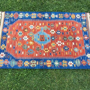Puede incluir: Una alfombra colorida con un patr&oacute;n geom&eacute;trico en tonos de rojo, azul, amarillo y blanco. La alfombra tiene un borde de flecos y est&aacute; hecha de lana.