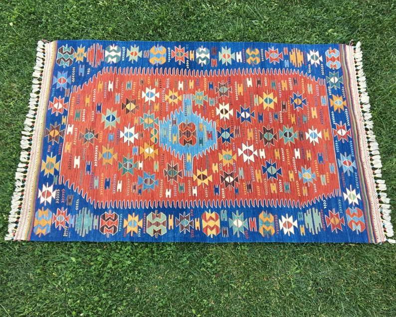Puede incluir: Una alfombra colorida con un patr&oacute;n geom&eacute;trico en tonos de rojo, azul, amarillo y blanco. La alfombra tiene un borde de flecos y est&aacute; hecha de lana.