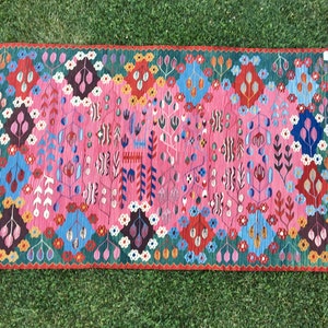 May include: A colourful rug with a pink background and intricate floral patterns in shades of blue, green, red, and white. The rug has a fringed border.