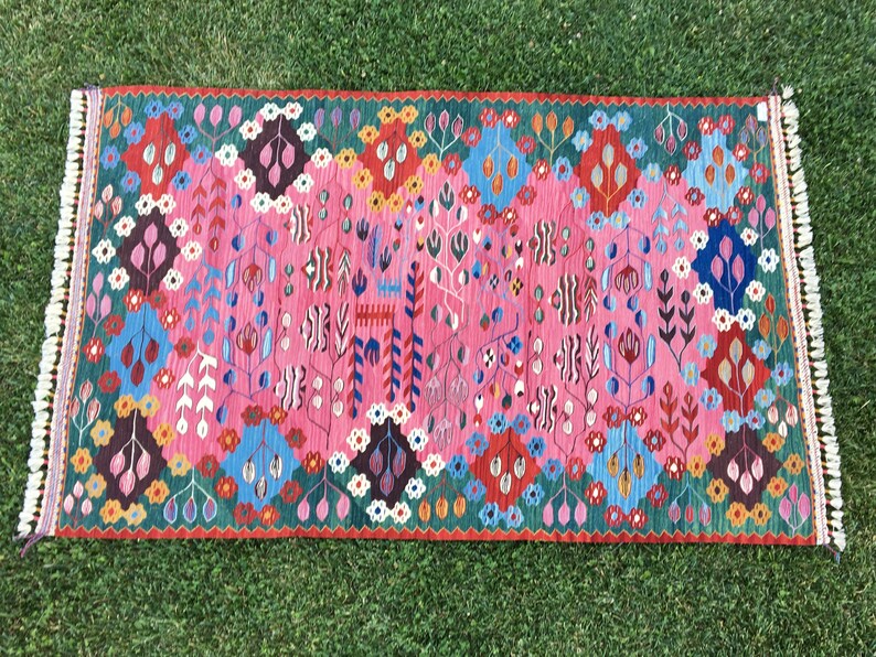 May include: A colourful rug with a pink background and intricate floral patterns in shades of blue, green, red, and white. The rug has a fringed border.