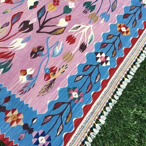 Op de afbeelding: Een kleurrijk tapijt met een geometrisch patroon met bloemen en bladeren. Het tapijt heeft een franje rand en is gemaakt van geweven stof.