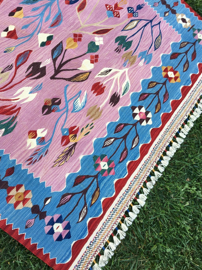 Op de afbeelding: Een kleurrijk tapijt met een geometrisch patroon met bloemen en bladeren. Het tapijt heeft een franje rand en is gemaakt van geweven stof.
