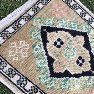 May include: A small, beige rug with a geometric pattern in shades of green, black, and pink. The rug has a white fringed border.