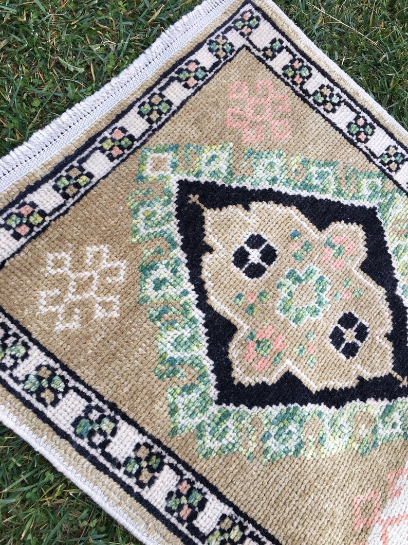 May include: A small, beige rug with a geometric pattern in shades of green, black, and pink. The rug has a white fringed border.