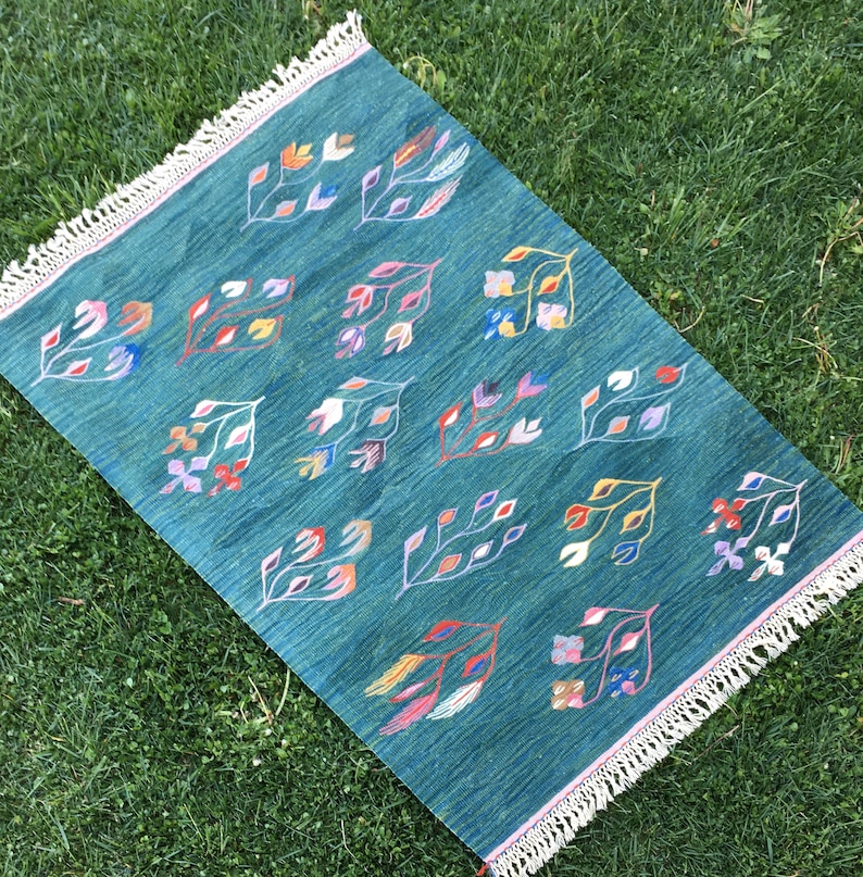 Puede incluir: Una alfombra azul turquesa con borde de flecos presenta un patr&oacute;n repetitivo de dise&ntilde;os florales estilizados de colores.