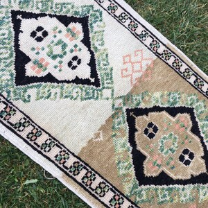 May include: A white and beige rug with a green, black, and pink geometric pattern. The rug has a fringed border.