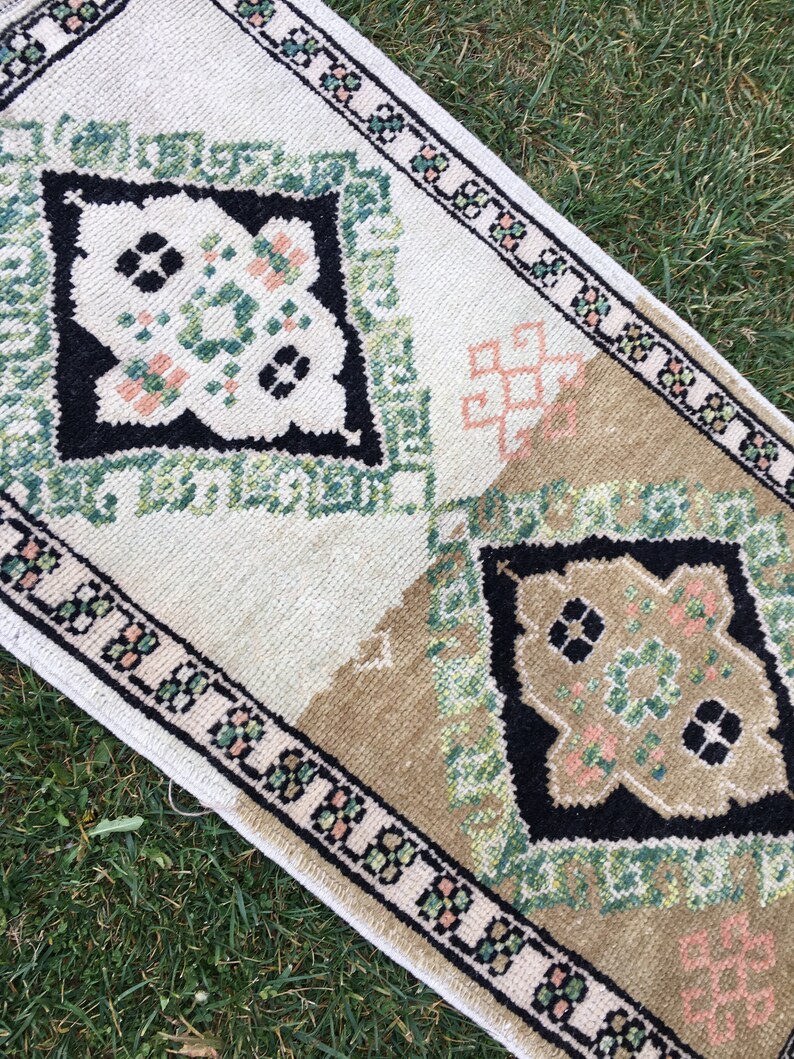 May include: A white and beige rug with a green, black, and pink geometric pattern. The rug has a fringed border.