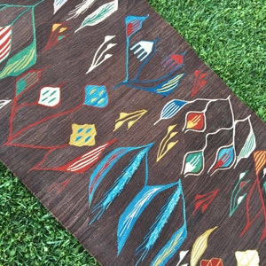 Puede incluir: Una alfombra marr&oacute;n con un patr&oacute;n geom&eacute;trico de hojas y flores de colores. La alfombra tiene un fondo marr&oacute;n oscuro con acentos blancos, azules, rojos, amarillos y verdes.