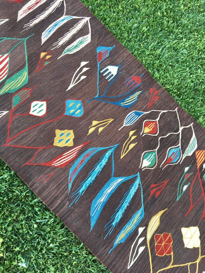 Puede incluir: Una alfombra marr&oacute;n con un patr&oacute;n geom&eacute;trico de hojas y flores de colores. La alfombra tiene un fondo marr&oacute;n oscuro con acentos blancos, azules, rojos, amarillos y verdes.