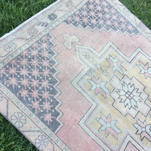 May include: A rectangular, vintage-style rug with a faded, distressed appearance. The rug features a central design in shades of yellow, pink, and gray, with a border in a darker gray. The rug is placed on a bed of green grass.