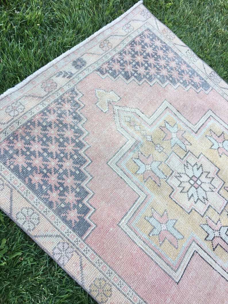 May include: A rectangular, vintage-style rug with a faded, distressed appearance. The rug features a central design in shades of yellow, pink, and gray, with a border in a darker gray. The rug is placed on a bed of green grass.