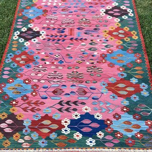 May include: A colourful rug with a pink background and a geometric pattern featuring flowers, leaves, and abstract shapes. The rug has a fringed border.