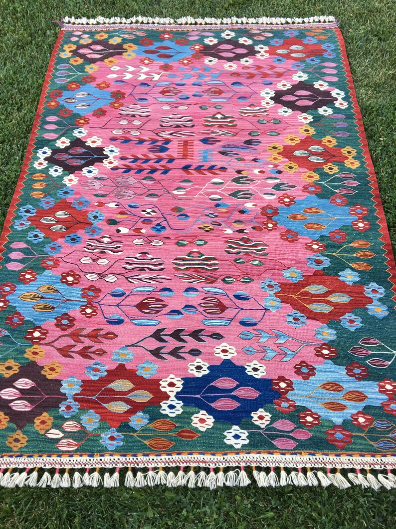 May include: A colourful rug with a pink background and a geometric pattern featuring flowers, leaves, and abstract shapes. The rug has a fringed border.