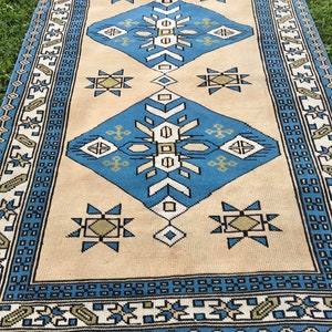 May include: A vintage rug with a cream background and blue and green geometric patterns. The rug has a fringe border.