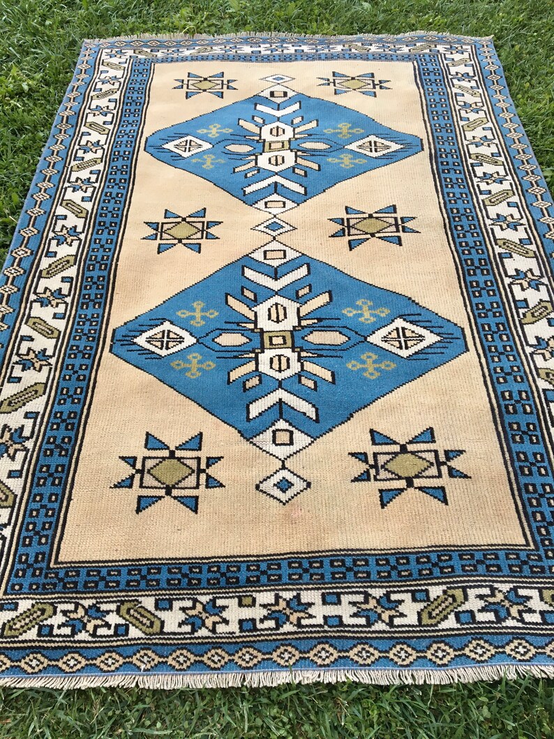 May include: A vintage rug with a cream background and blue and green geometric patterns. The rug has a fringe border.
